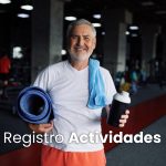 Hombre mayor sonriente en un gimnasio, sostiene una esterilla de ejercicio y una botella de agua, con una toalla azul alrededor del cuello. Fondo con equipos de ejercicio.