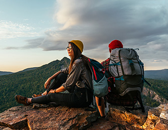 Pareja sentada en una roca, mirando el paisaje montañoso al atardecer, con mochilas y ropa de senderismo.