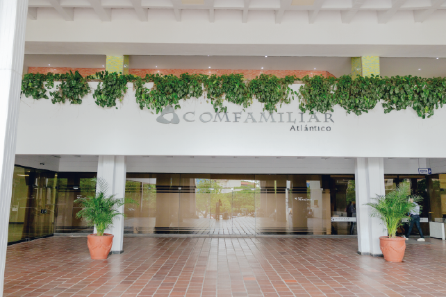 Entrada principal de un edificio con letrero "Comfamiliar Atlántico", plantas decorativas en macetas y un ambiente luminoso. Suelo de cerámica y paredes blancas.
