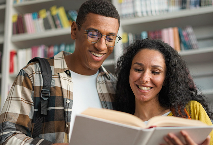 Dos personas sonrientes observan un libro en una biblioteca, rodeadas de estanterías con libros de diferentes colores. El ambiente es iluminado y acogedor.