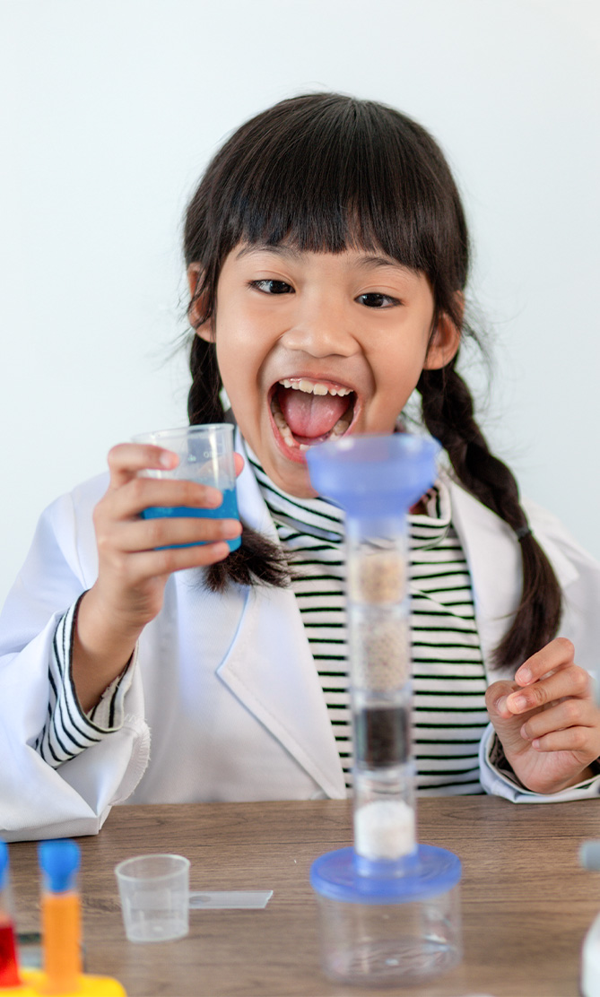 Niña con bata de laboratorio sostiene un vaso con líquido azul, sonriente, frente a un cilindro graduado con capas de sustancias. Fondo blanco y mesa de trabajo con material de laboratorio.