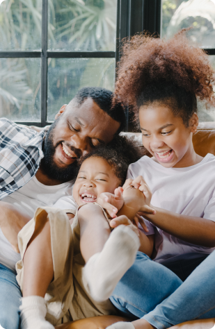 Hombre adulto y dos niños se ríen y juegan sentados en un sofá, rodeados de luz natural. Los niños tienen cabello rizado y llevan ropa casual.