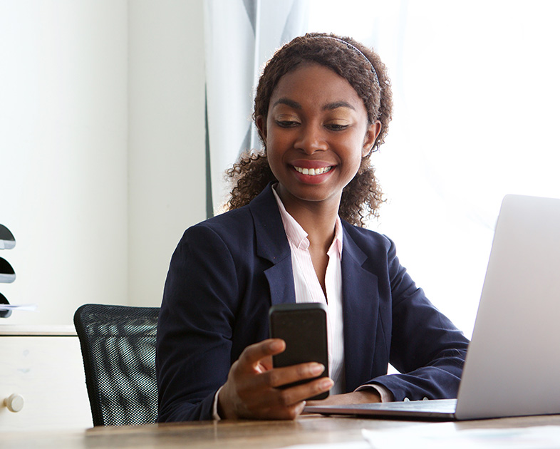 Mujer sonriente revisando el teléfono móvil, sentada frente a una computadora portátil en un entorno de oficina.