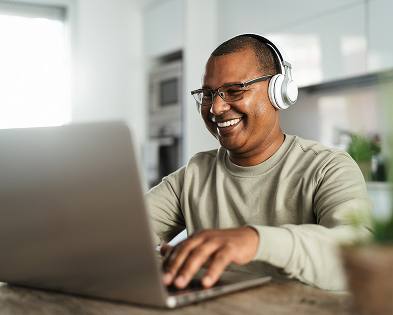 Hombre con auriculares sonriente, usando una laptop en una mesa de madera. Entorno luminoso y moderno, con plantas y muebles de cocina al fondo.