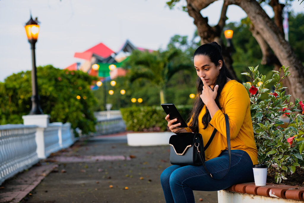 Mujer sentada en un banco, mirando su teléfono móvil, con un entorno de jardines iluminados y edificios coloridos al fondo.