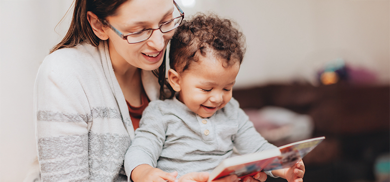 Mujer con gafas y niño pequeño sentados juntos, leyendo un libro. Ambos sonríen, en un entorno acogedor con colores suaves y desenfocados al fondo.
