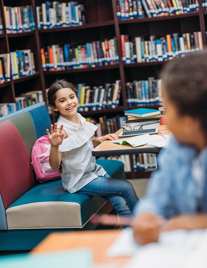 Niña sonriente con blusa blanca y mochila rosa, sentada en una biblioteca, saludando mientras estudia con libros y cuadernos sobre la mesa. Fondo con estanterías llenas de libros.