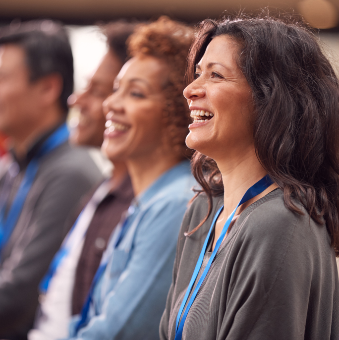Grupo de personas sonriendo y escuchando, con collares de identificación. Predominan tonos de piel diversos y ropa en colores neutros y azules, en un entorno de evento o reunión.